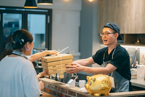 A pizza shop owner personally hands over a freshly baked and well-packaged pizza to the customer, ready for them to enjoy the delicious meal.
