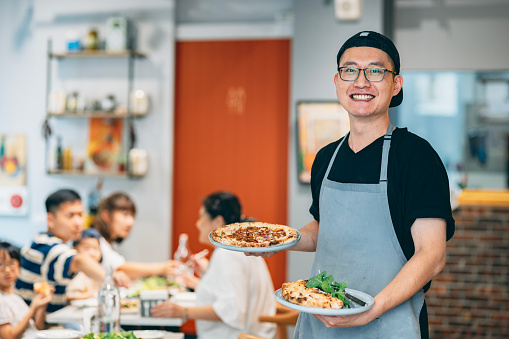 This is a portrait photo of an Asian chef standing in a restaurant, holding the pizza he has baked. In the background, there are customers enjoying their food. He looks at the camera with a confident smile.