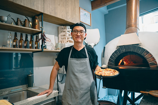 This is a portrait photograph capturing an Asian chef who skillfully crafts delicious pizzas. He exudes confidence as he holds a freshly baked pizza and gazes directly at the camera.