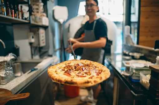 An Asian pizza chef uses a pizza peel to carefully remove the freshly baked pizza from the oven. He is preparing to place it on a plate and serve it to the customers for them to enjoy.The focus is on the pizza, and the chef looks directly at the camera.