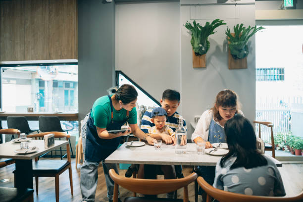 An Asian female restaurant staff member is holding a tablet computer to record the customer's order while engaging in friendly interaction, providing attentive table service. una atenta empleada asiática en una pizzería está proporcionando servicio de mesa a un cliente. se agacha cálidamente a la altura de los ojos con el cliente, estableciendo una conexión amistosa, y presenta con entusiasmo los platos destacados del rest
