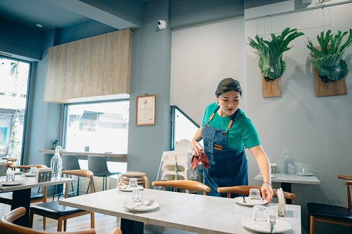 Before the restaurant's opening hours, an Asian female staff member in a pizza shop is cleaning the tables and arranging items such as menus, cutlery, and glasses in preparation for the arrival of customers.