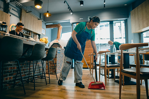 Before the restaurant's opening hours, the owner is at the cash register, verifying today's meal orders and cross-checking the ordered ingredients with the amounts. Meanwhile, the staff members are cleaning the surroundings.