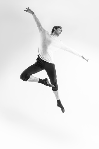 Professional Caucasian Handsome Young Athlete Man Posing in Flying Ballet Pose with Lifted Hands in White Shirt On White.Black and White Image