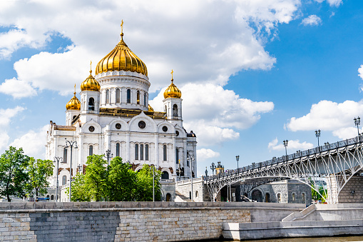 Cathedral of Christ the Saviour and Patriarshy Bridge, situated along the Moskva River in Moscow, Russia. The Cathedral of Christ the Saviour, known as Khram Khristá Spasítelya, stands prominently against a backdrop of blue sky with fluffy clouds.