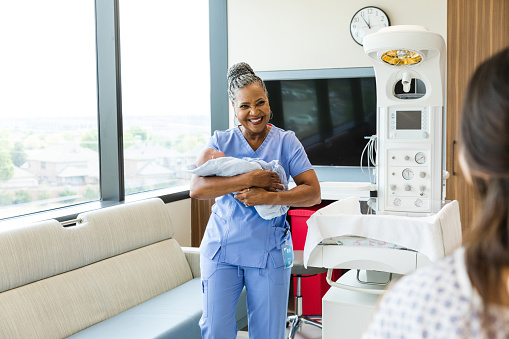 The smiling senior adult female doctor carries the newborn to the unrecognizable new mom.