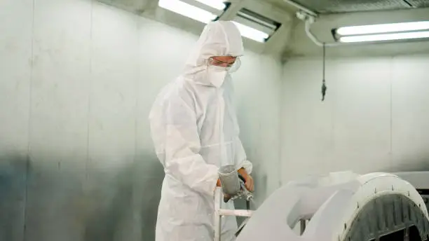 Working in an anti-wind car spray booth Of a Caucasian man in a white PPE protective suit spraying primer on a car bumper. sent to fix the paint in the car repair with intention and meticulousness