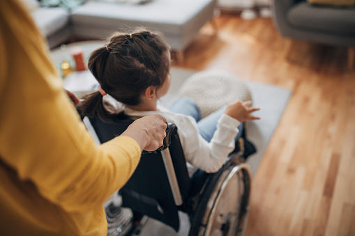Mother pushing her young daughter who is in wheelchair in living room at home.