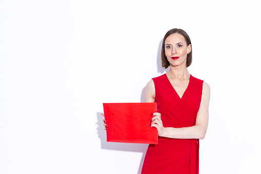 Sensual Portrait of Winsome Positive Confident Caucasian Business Woman in Long Red Dress Demonstrating Red Folder Over White Background. Horizontal Image