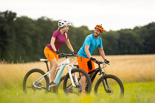 happy couple cycling electric mountain bikes through high dry grass in rural lanscape in summer smiling at each other