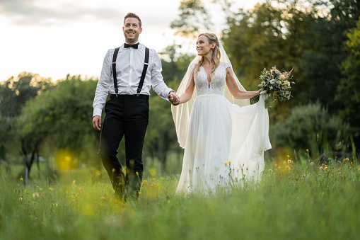 beautiful happy smiling romantic wedding couple walking together hand in hand through summer meadow at sunset, bride with bridal bouquete looking at her husband