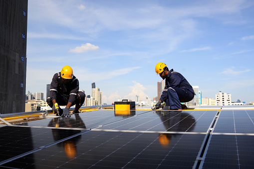 Male technicians carrying photovoltaic solar module on roof top of factory. Engineers in helmets installing solar panel system outdoors. Concept of alternative and renewable energy.