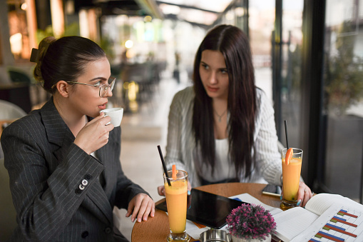 Two Businesswomen Discussing Economics While Drinking Coffee And Fresh Orange Juice