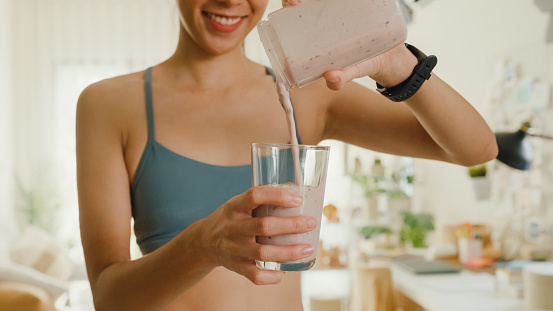 Closeup of young Asian athletic woman using blender and pouring smoothie to glass at home. Healthy food concept.