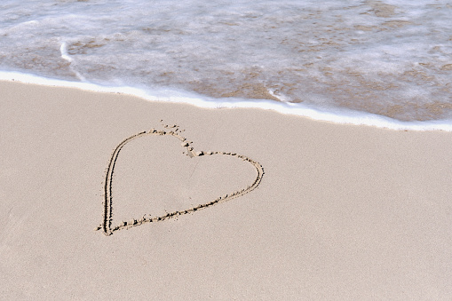 Wave running to heart shape drawn on white sand of tropical beach. Copy space