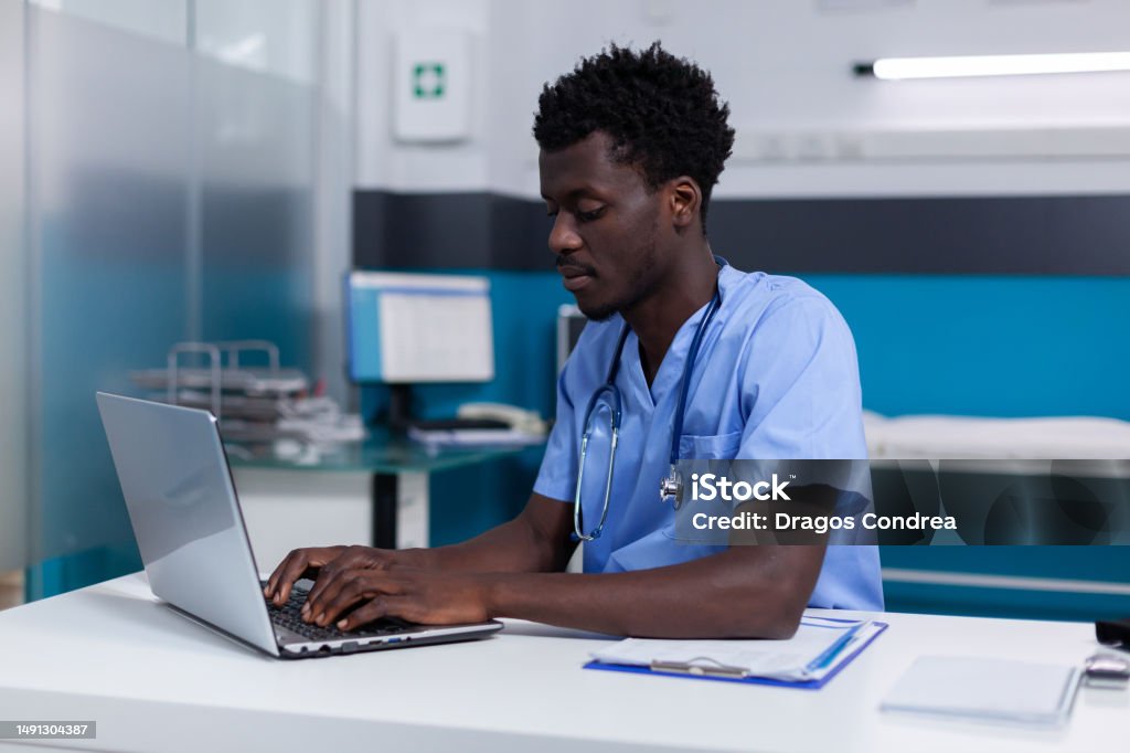 Black young man working as nurse at medical clinic Black young man working as nurse at medical clinic wearing uniform and stethoscope. African american person sitting at desk while using modern laptop, typing on keyboard in cabinet Nurse Stock Photo
