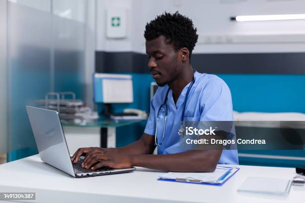 Black Young Man Working As Nurse At Medical Clinic Stock Photo - Download Image Now - Nurse, Typing, Cabinet