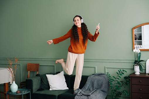 Joyful young woman dancing to her favorite music at home. Happy girl experiencing joy while listening to popular music with headphones. Pretty girl having fun and dancing to music in her living room.