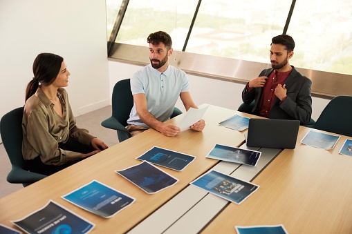 Captured from a high angle, three businesspersons gather around a table filled with scattered technology devices and business charts.