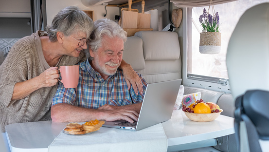 Happy carefree senior couple in travel vacation leisure sitting inside a camper van dinette enjoying breakfast together while using laptop
