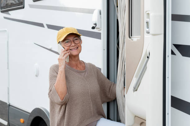 happy senior woman standing outside a camper van motor home talking on mobile phone enjoying travel destination. smiling elderly lady in alternative vacation transport and tourism concept - motor home mobile home camping senior adult imagens e fotografias de stock