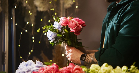 Close up shot of male florist entrepreneur preparing bouquet for sale or delivery. Flower shop worker collects bunch of beautiful flowers. Vases with plants at background. Concept of floral business.