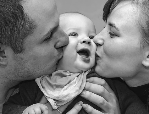 Portrait of a young caucasian family, a young man and woman hug and kiss a little baby. Black and white photo.