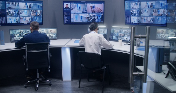 Diverse team of officers watch CCTV cameras with AI facial recognition. Employees work in security control center. Computer monitors and big digital screens showing surveillance cameras video footage.