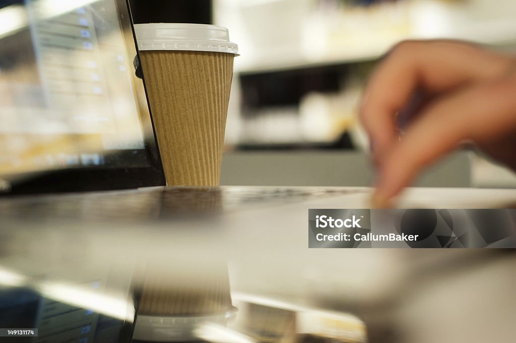 Disposable Coffee cup by working laptop In sharp focus is a disposable coffee cup slightly behind a laptop. In the foreground and very blurred, is a laptop being worked on (visible hand). This is in a meeting environment. Business Stock Photo