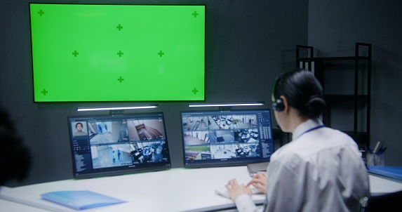 Female officer controls CCTV cameras with AI facial recognition in security control center with colleague. Computer monitors showing surveillance cameras footage. Big digital screen with chroma key.