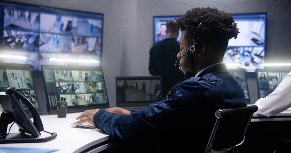 Multi ethnic employees talk and control CCTV cameras with AI face scanning in surveillance center together. Multiple big screens on the wall showing security cameras footage. Concept of social safety.