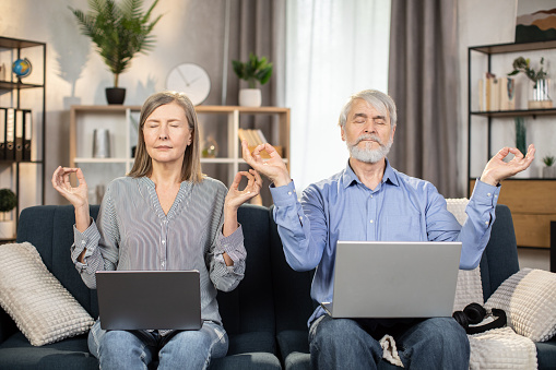Portrait of aged spouses holding fingers in gyan mudra while relaxing on couch with laptops on knees in room interior. Gray-haired man and woman reducing fatigue using meditation practice at home.