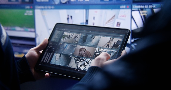 Close up shot of operator zooming and monitoring CCTV cameras using tablet in police surveillance center. Big screen on the wall showing security cameras with AI face scanning footage. Social safety.