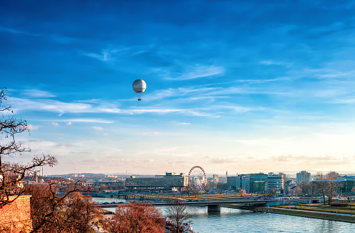 Krakow city over Vistula river with flying hot air balloon and Ferris wheel in Poland. Autumn cityscape with river walking people and ferris wheel in distance