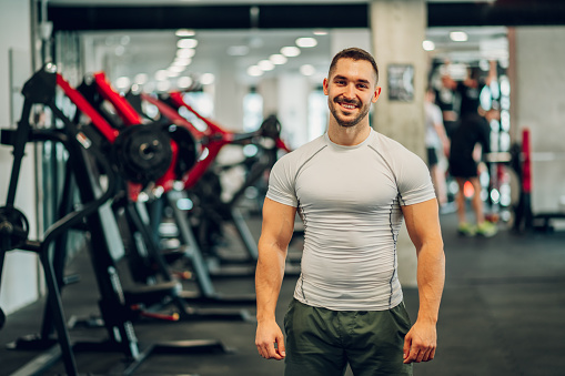 A cheerful healthy sportsman is posing in a gym and smiling at the camera. A happy bodybuilder in activewear is standing at a sports center with exercise equipment in a blurry background.