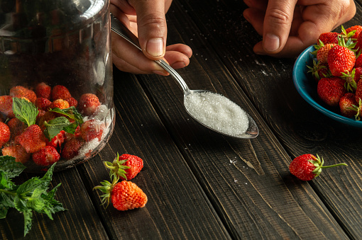 Hand of cook with spoon on kitchen table. Sugar is added to a jar of ripe strawberry and mint compote