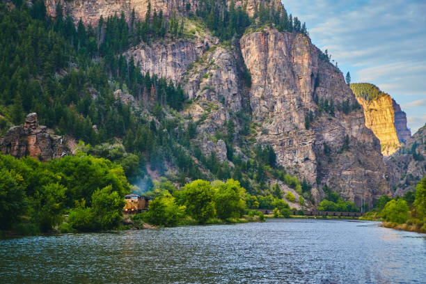 istock Train on tracks by river at bottom of large canyon with rock cliffs and pine trees 1491322671
