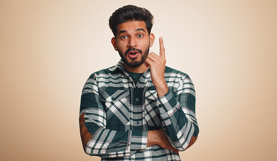 Eureka. Inspired young indian man pointing finger up with open mouth, having good idea, plan, startup, showing inspiration motivation gesture, problem solution. Bearded hindu guy on beige background