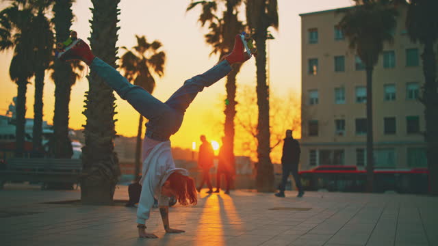 SLO MO Young woman in roller skates performs acrobatic moves in a square surrounded by palm trees at sunset