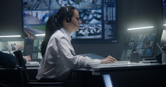 Female operator in headset types keyboard and controls security cameras with AI face scanning. PC monitors and big digital screens on the wall show CCTV cameras footage. Work in surveillance center.