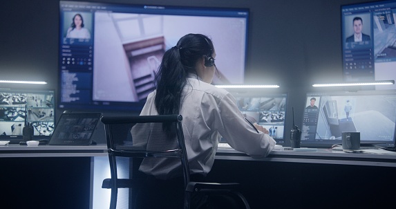 Female operator types keyboard and monitors security cameras with AI facial recognition in police surveillance room. PC monitors and big digital screens on the wall with displayed CCTV cameras view.