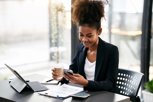 African american businesswoman in suit holding coffee cup and reading accounting information of new business on smartphone while working on tablet with paperwork in modern workspace.