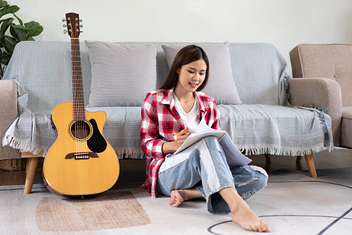 Young woman is compose the song and writing lyrics on notebook after playing guitar while sitting on the floor in living room at home.