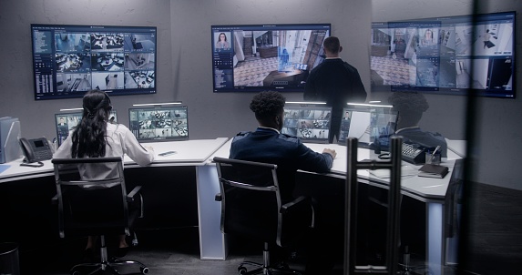 Multi ethnic security officers monitor CCTV cameras with AI face scanning. Multiple big screens with displayed security cameras. Teamwork in surveillance room. Monitoring, tracking and social safety.