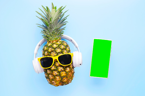 Fancy pineapple in sunglasses and wireless headphones listening to music with phone on a blue background