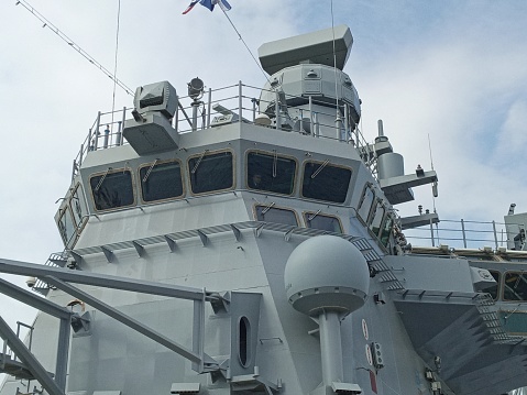 One crew member is raising two maritime flags, while the other watches or assists, on the bridge of USS Canberra (LCS 30), an Independence Class combat ship of the US Navy which is docked at Garden Island in Sydney Harbour.  She was in port for her commissioning ceremony on 22 July 2023.  As part of the ceremony, her captain was presented with a kangaroo insignia attached to all ships of the Royal Australian Navy, but decorated in the colours and design of the Stars and Stripes.  That insignia has been attached to the ship and visible on the right.  The name of the ship and serial number is visible on the orange life ring.  A large, padded chair is visible on the bridge.  This image was taken on a cloudy afternoon on 29 July 2023.