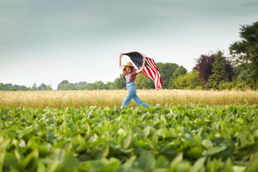 Subject: A young farm girl running across the farm field with United States flag flying behind her.