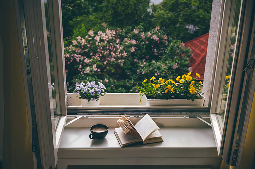 Open book and cup of coffee on windowsill in front of the open window. Home comfort zone. Summer time