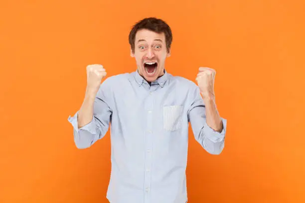Portrait of extremely happy man standing with clenched fists, celebrating his success, screaming hurray, wearing light blue shirt. Indoor studio shot isolated on orange background.