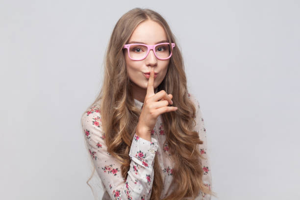 Woman showing shh gesture holding finger near lips, with naughty smile on face, making surprise. Portrait of woman in glasses with wavy blond hair showing shh gesture holding finger near lips, with naughty smile on face, making surprise, secret. Indoor studio shot isolated on gray background. n11 stock pictures, royalty-free photos & images